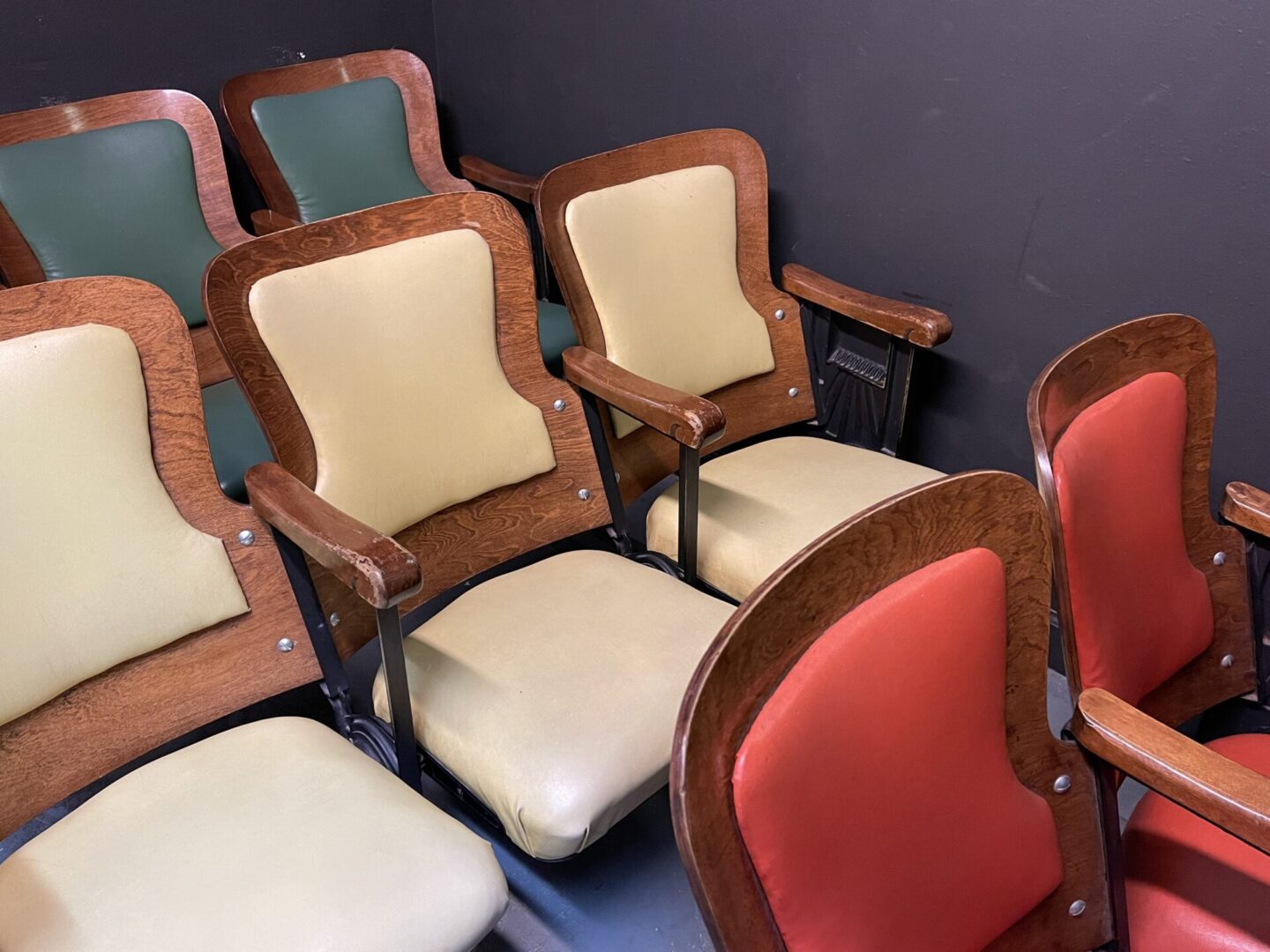 A row of vintage theater seats with wooden frames and multicolored cushions in red, green, and beige against a dark wall.