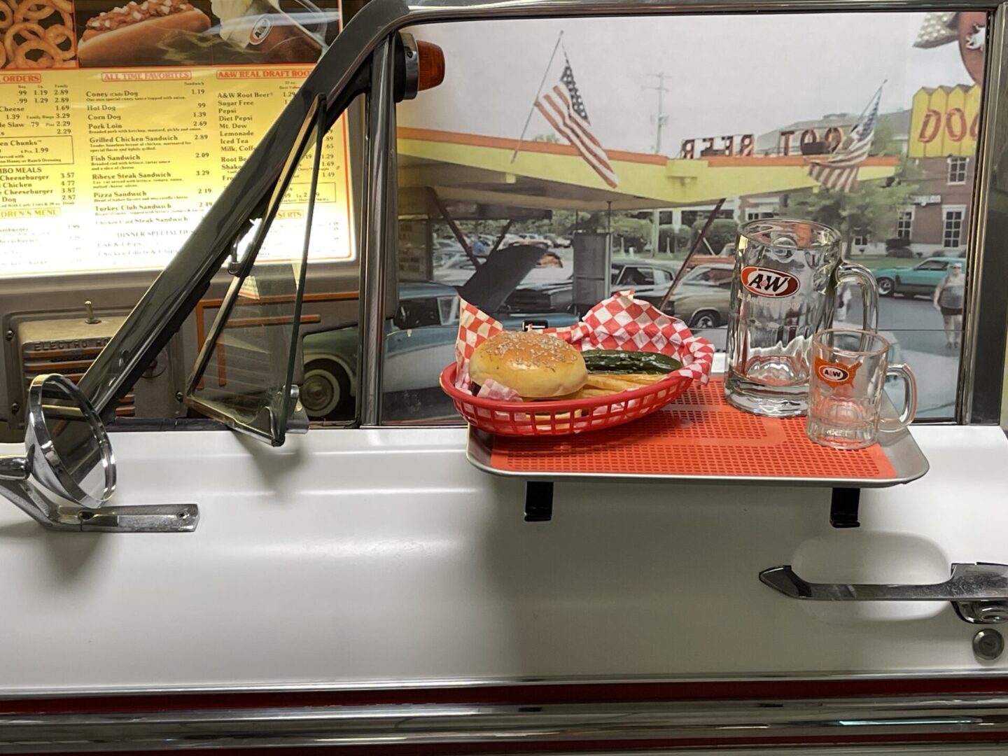 Car window tray with a cheeseburger, pickles, a glass jug, and an A&W mug at a drive-in diner with a vintage aesthetic.