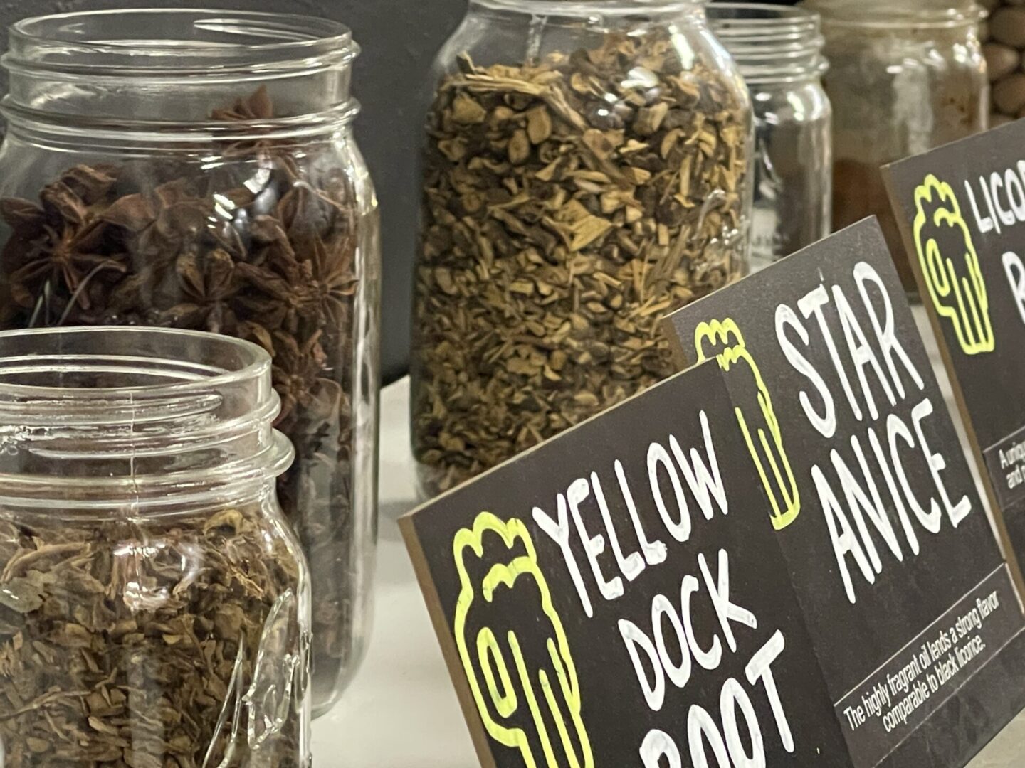 Jars filled with dried herbs including yellow dock root and star anise, labeled with black signs on a shelf.