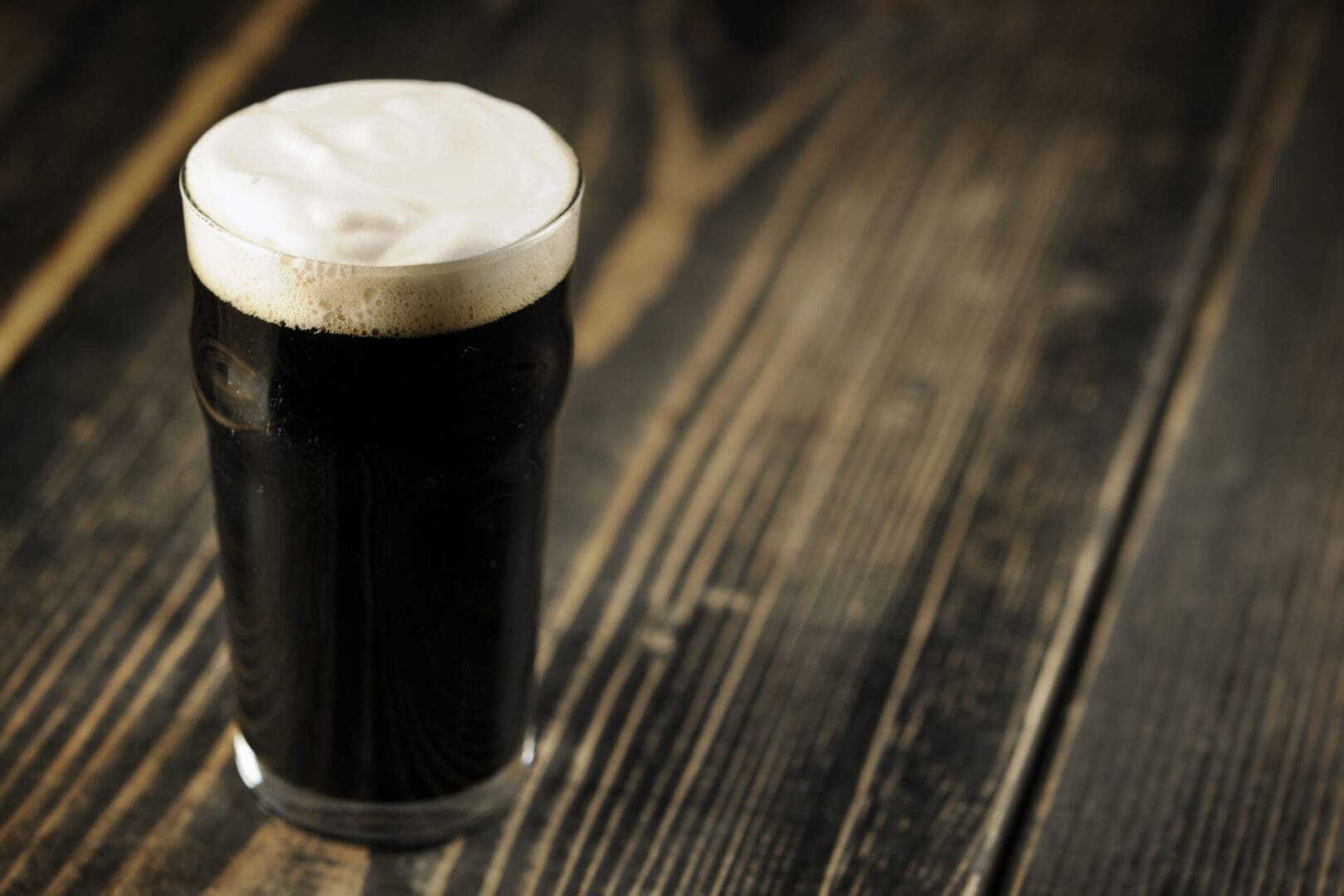 A glass of dark beer with a frothy head sits on a rustic wooden table.