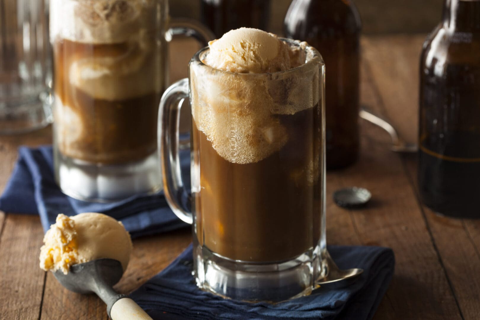 Two glass mugs filled with root beer float, topped with vanilla ice cream. A scoop of ice cream is placed beside the mugs on a wooden surface with a blue cloth.