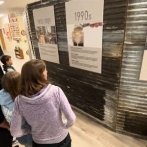 A group of people looking at a museum exhibit displaying historical information on a corrugated metal wall.