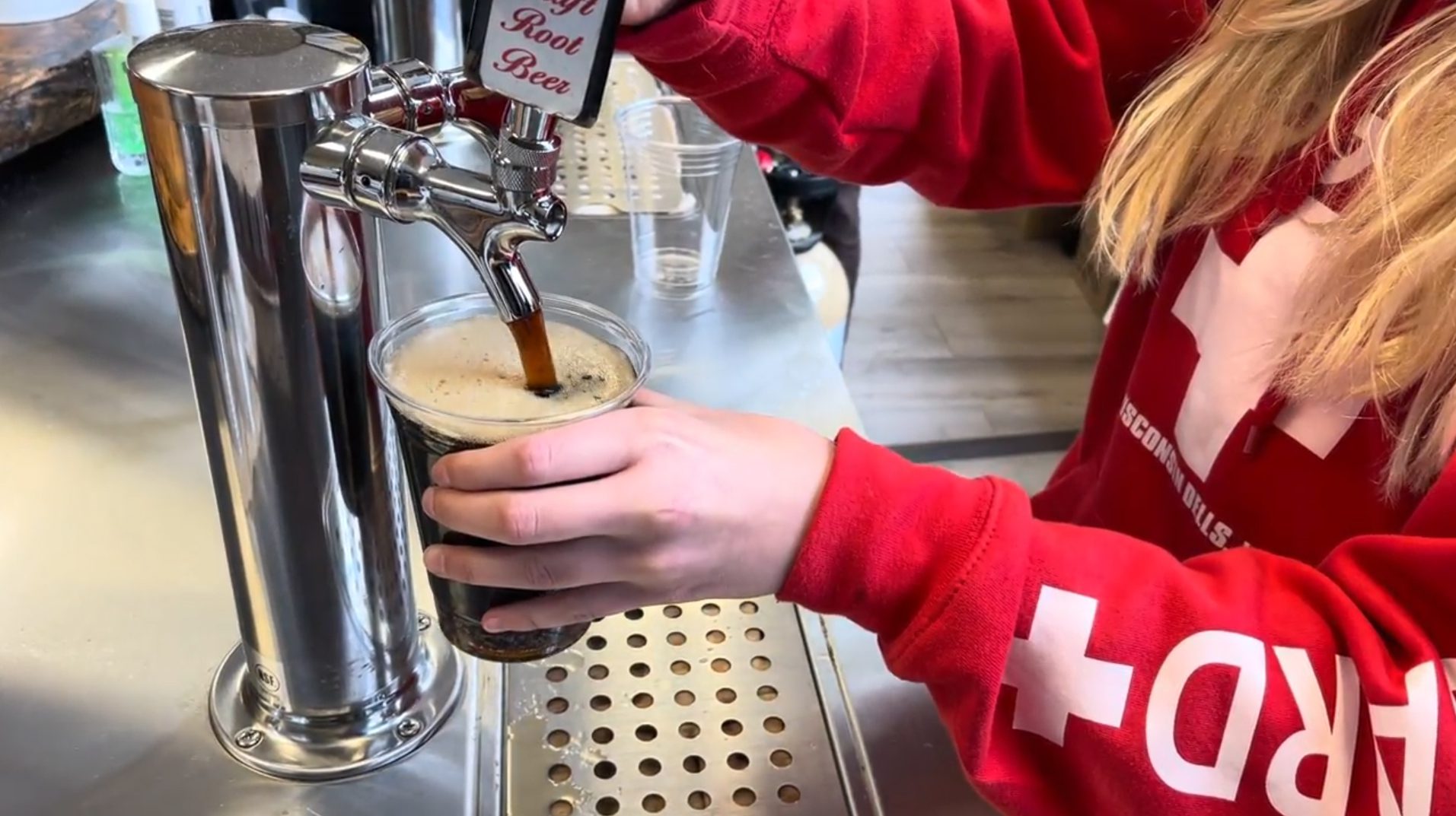 A person in a red hoodie fills a cup with root beer from a tap.