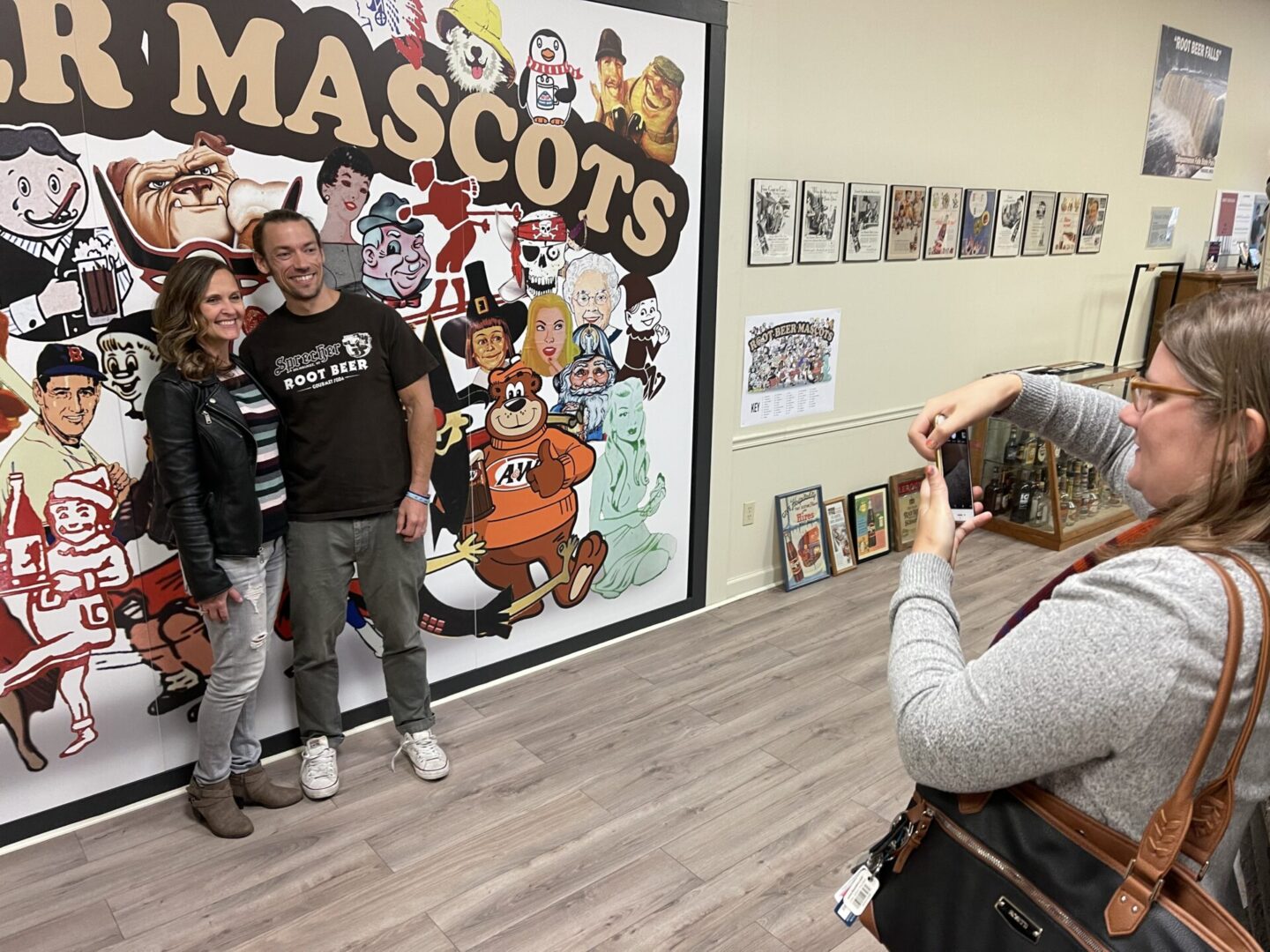 Two people pose in front of a cartoon mascots mural while another person takes their photo in a room with framed pictures on the wall.