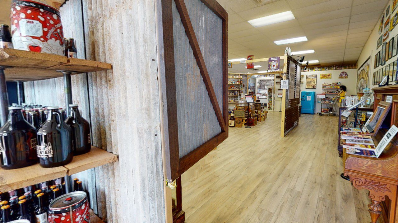 A rustic interior of a store with wooden shelves displaying jars and bottles. A large wooden door is partially open, leading to an aisle with more shelves and displays.