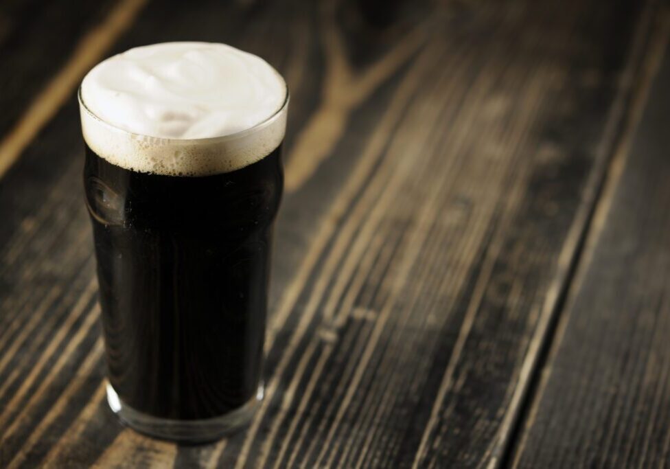 A glass of dark beer with a frothy head sits on a rustic wooden table.