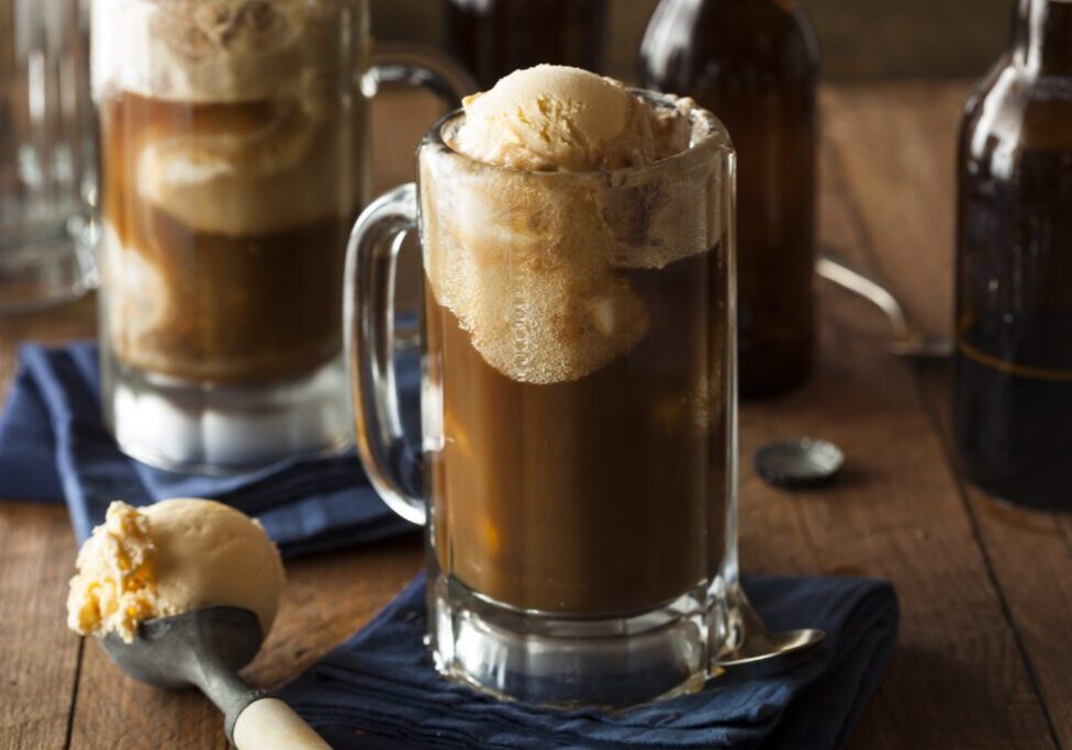 Two glass mugs filled with root beer float, topped with vanilla ice cream. A scoop of ice cream is placed beside the mugs on a wooden surface with a blue cloth.